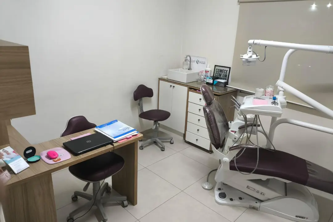 Sala de atendimento da clínica odontológica, mostrando a cadeira de dentista, bancada com pia, armários e mesa de planejamento com notebook.